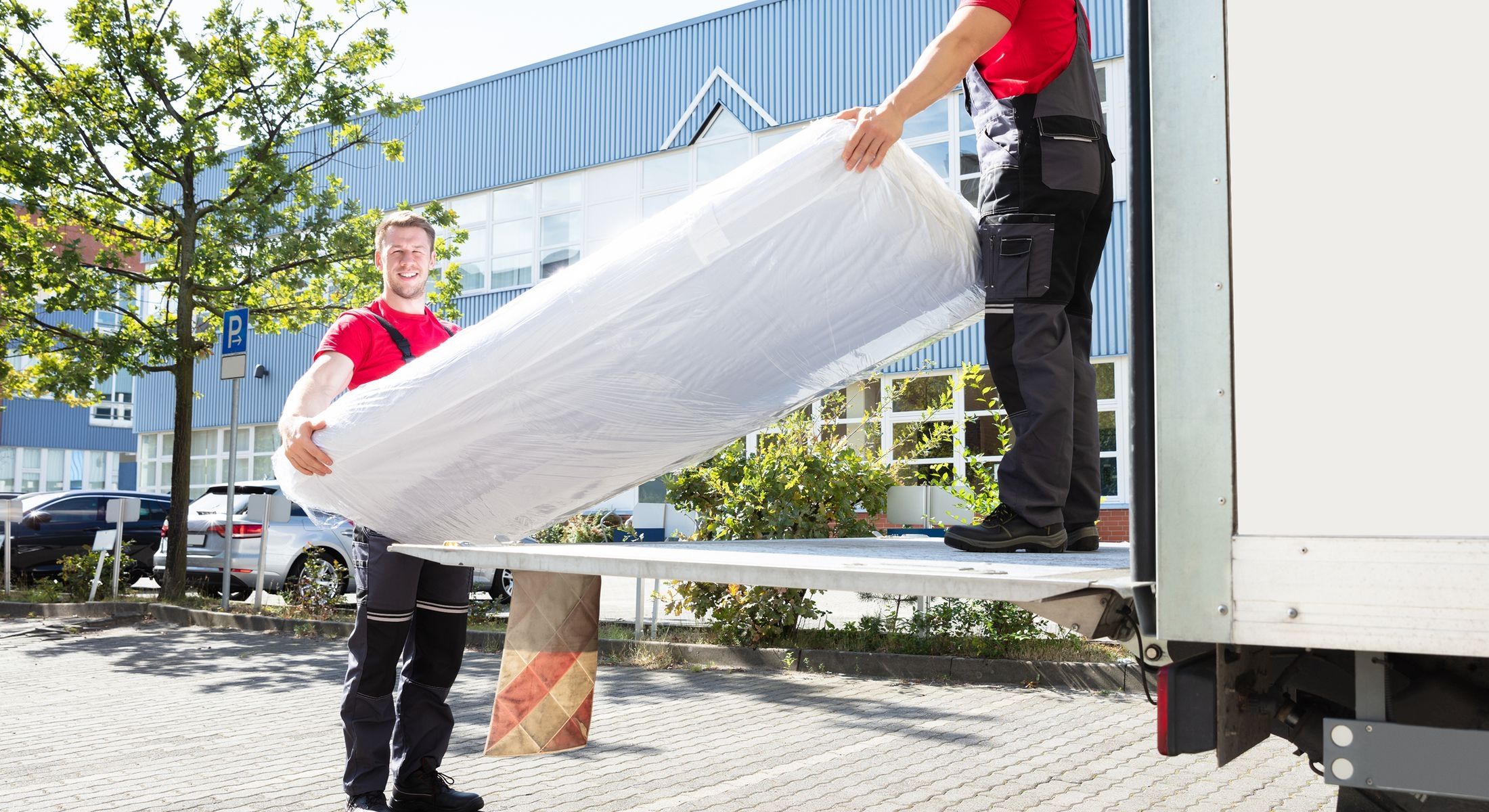 Zwei Möbelpacker in roten Shirts tragen eine in Schutzfolie verpackte Matratze aus einem Umzugswagen